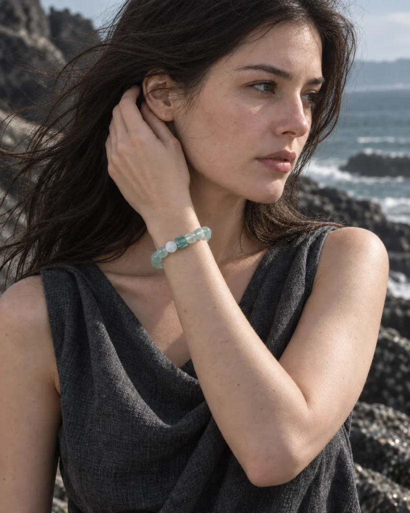 Model wearing the Tidal Glass green fluorite bracelet near her chin beside coastal rocks