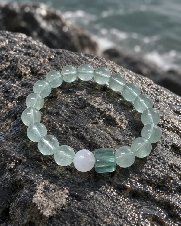 Tidal Glass green fluorite bracelet resting on a dark coastal rock beside water